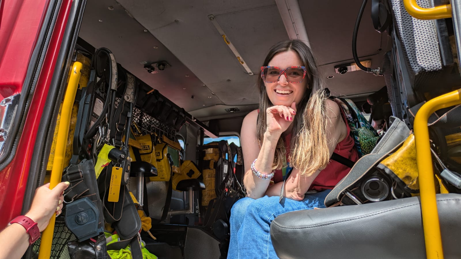 A young woman with long dark hair is wearing blue jeans and beige top. Wearing sunglasses she smiles as she sits in a fire engine.