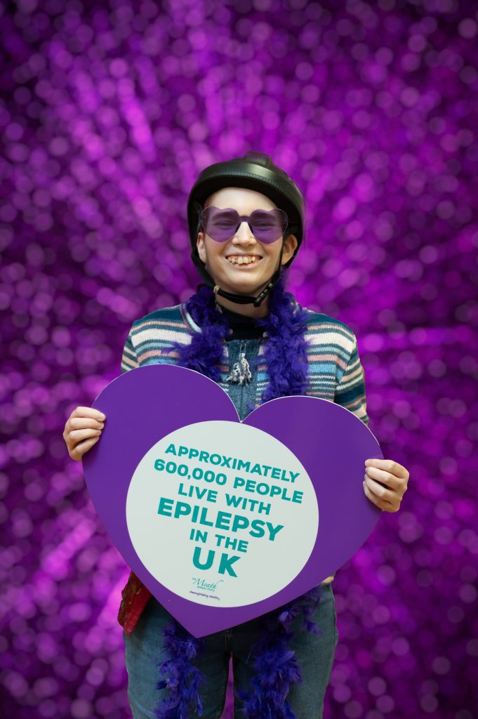 A woman wears a black epilepsy helmet and purple heart shaped glasses. she wears a purple feather boa and is stood up against a purple backdrop.