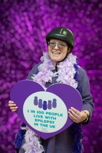Nancy in purple holding a cut our heart which reads '1 in 100 people in the UK have epilepsy'