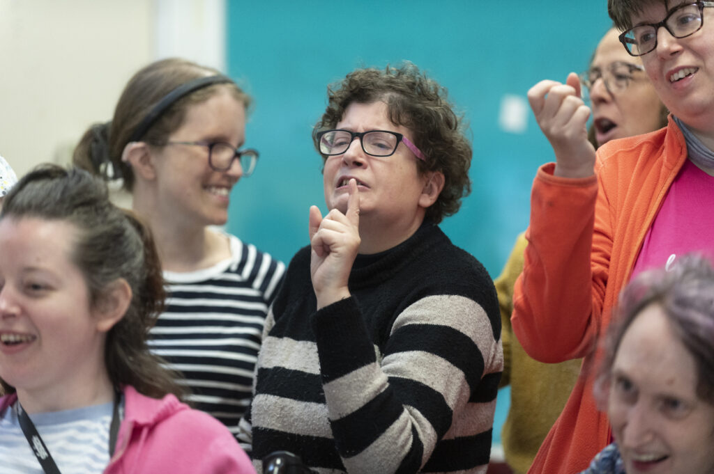 A woman wearing a stripy top is signing in Makaton. Next to her are two women who are smiling at her.