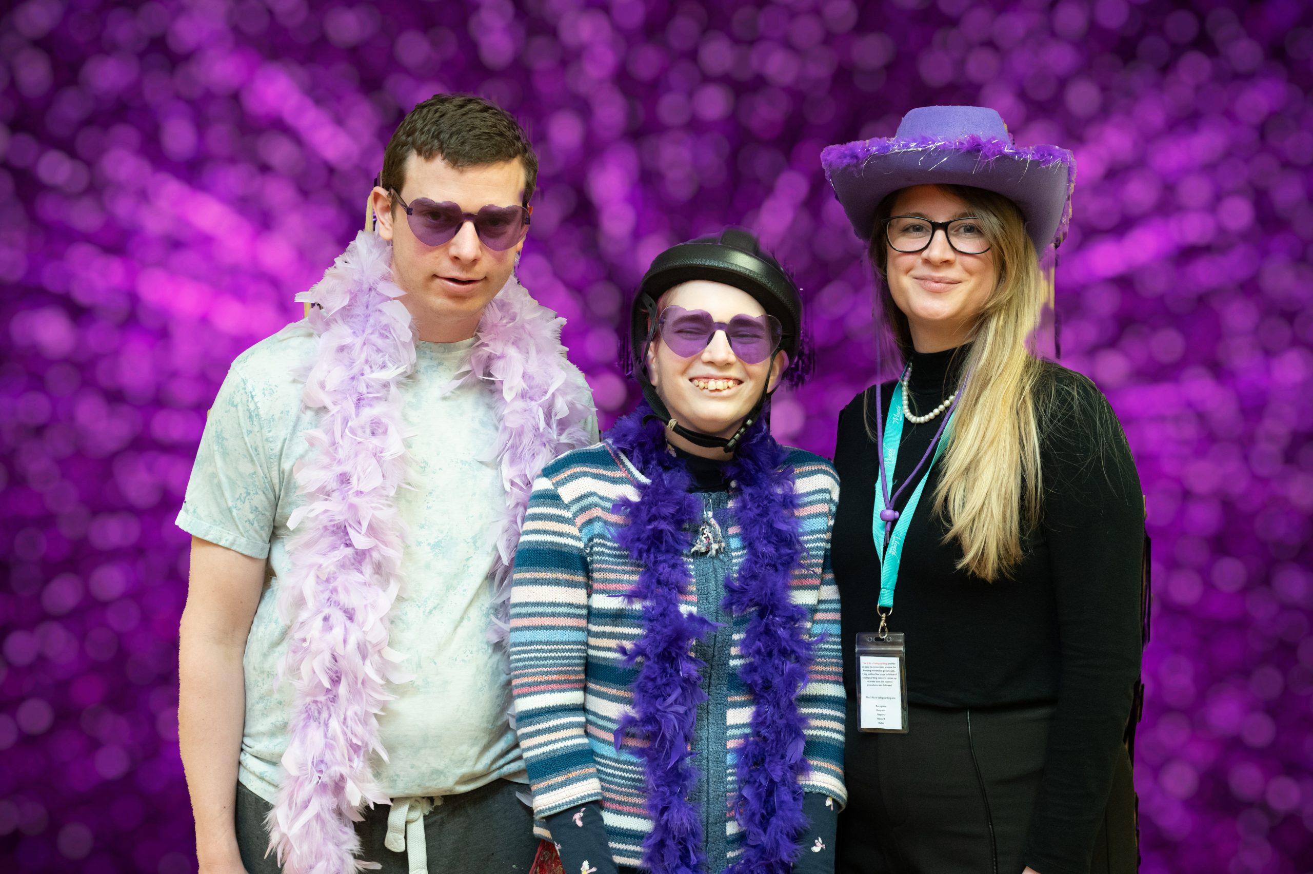 A man and two women wear lots of purple accessories and stand in front of a bright purple background as they smile to the camera.