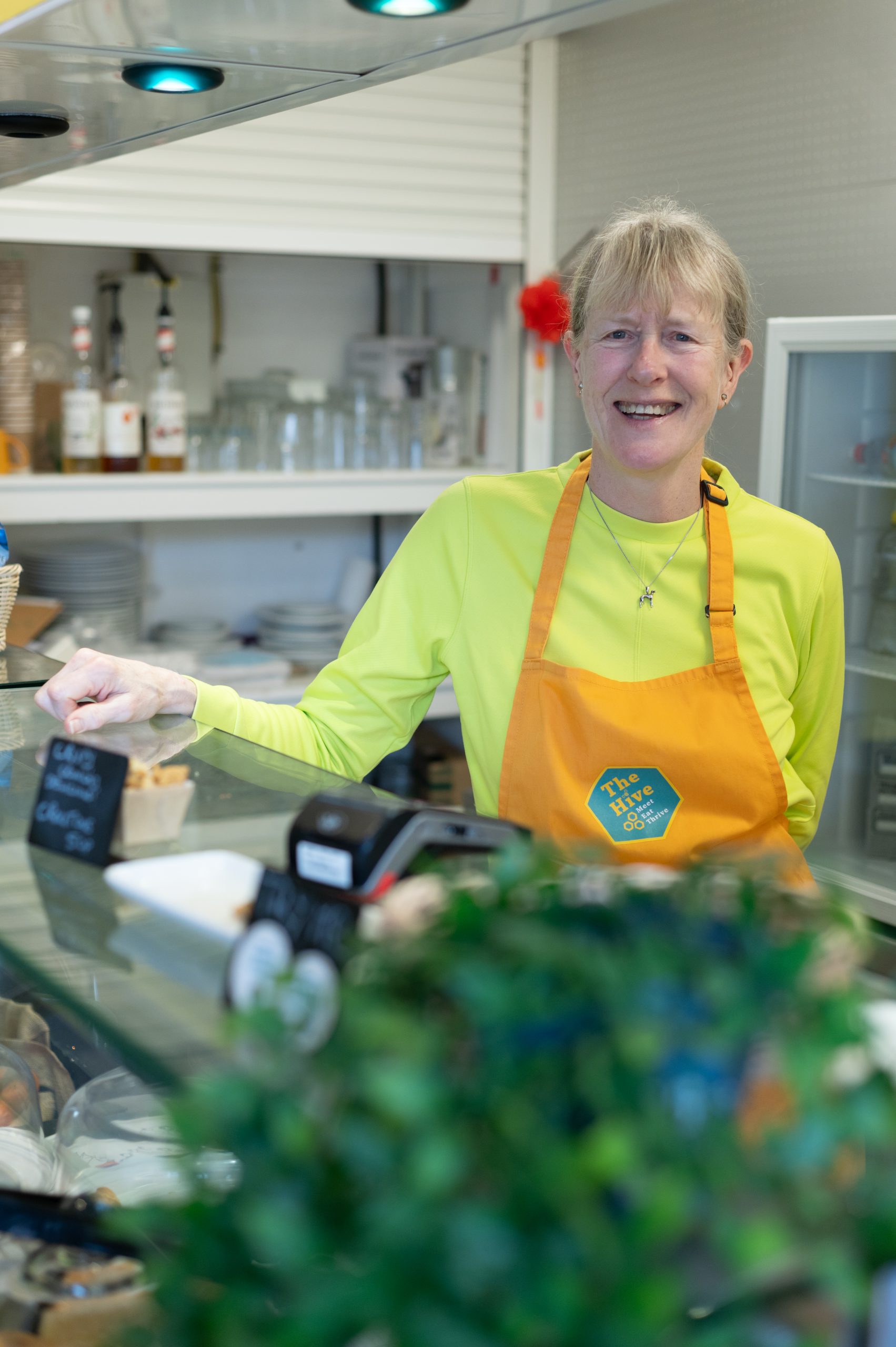 A woman in a yellow apron is stood behind a cafe counter and is smiling.