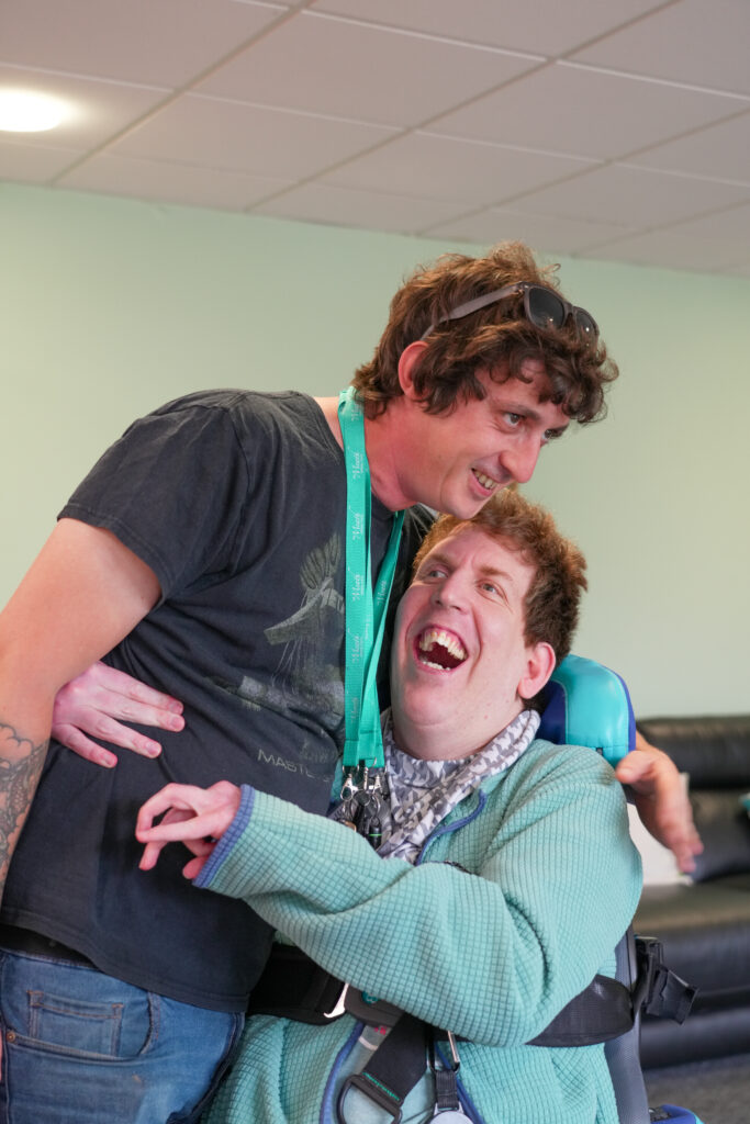 A male Support Worker is smiling while being hugged by a male person her supports.