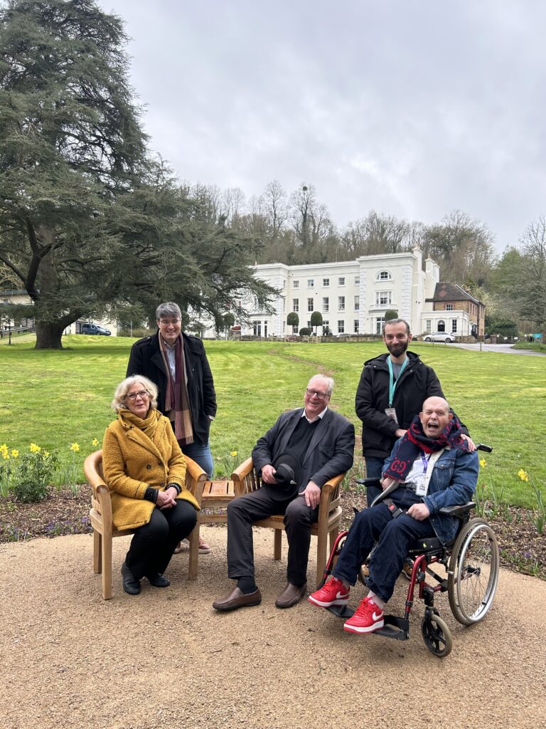 Cllr. Penny Rivers, Mayor of Godalming sits in the Woodland Walkway area at The Meath in a yellow coat. She is with Cllr. Paul Follows, a resident and their Support Worker.