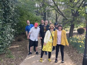 A group of men and women are stood on a woodland path and smiling the camera. A young man is giving a thumbs up to the camera. Around them are yellow daffodils. 