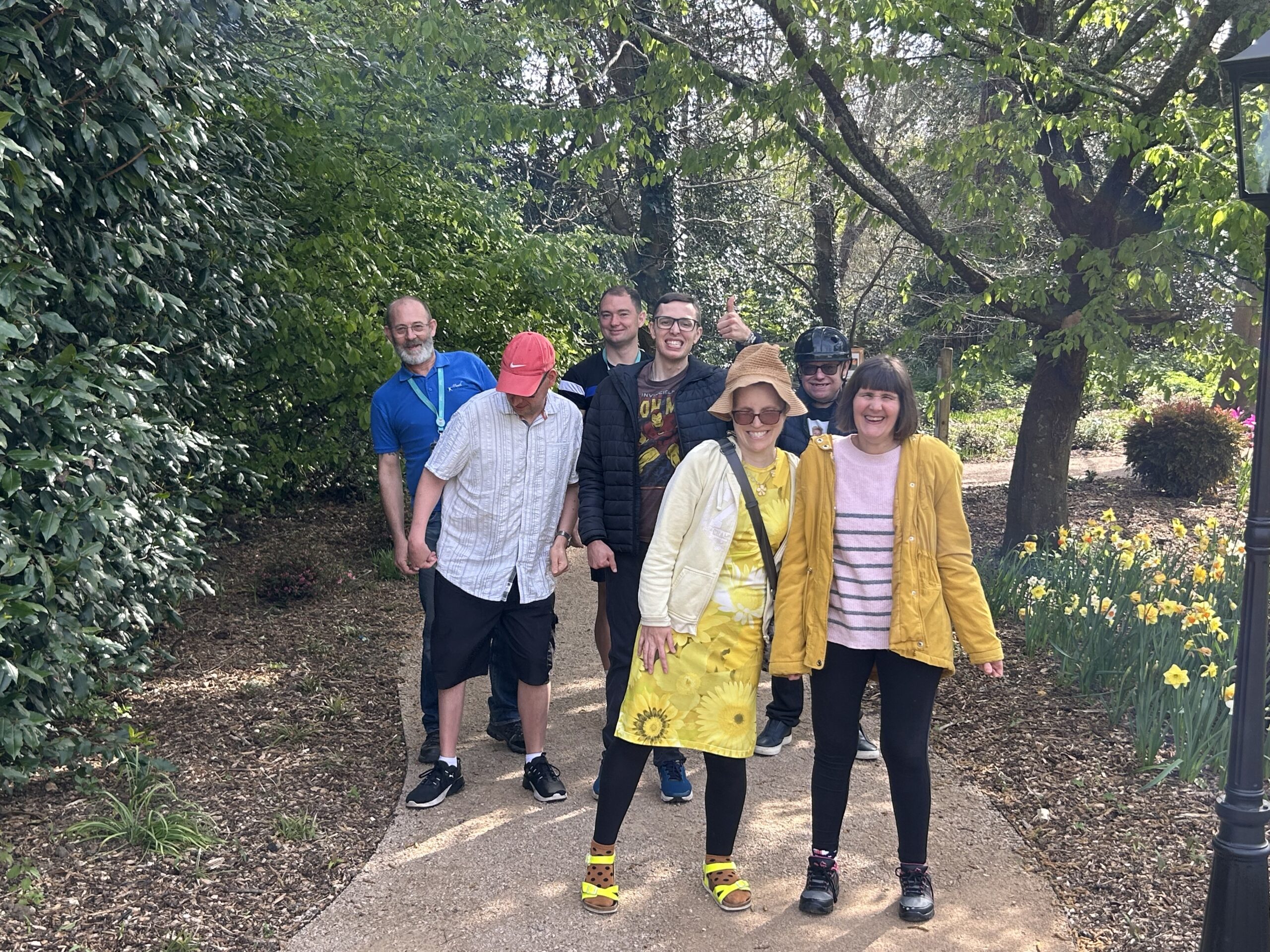 A group of men and women are stood on a woodland path and smiling the camera. A young man is giving a thumbs up to the camera. Around them are yellow daffodils.