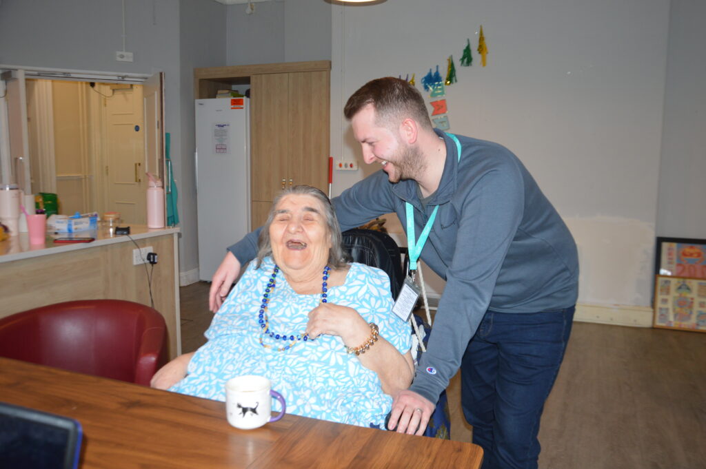 An elderly lady in a wheelchair is at her kitchen table with a cup of tea laughing with her support worker who is smiling at her.