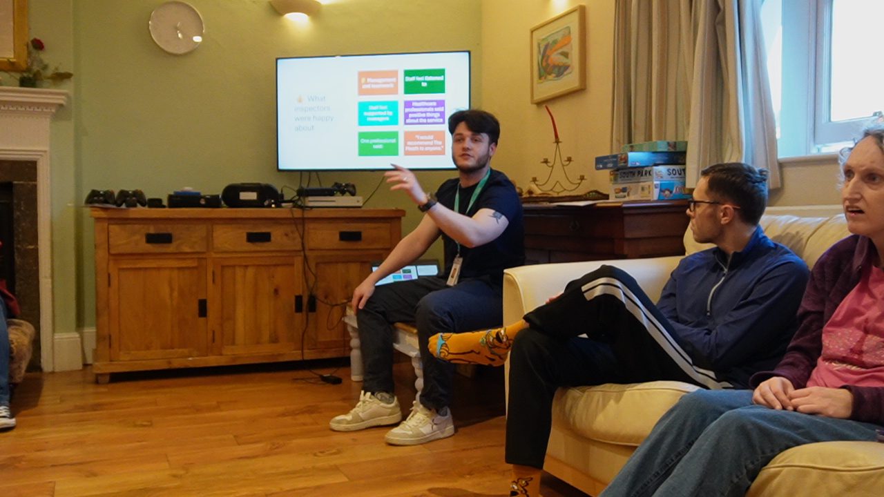 A man is talking as he explains a presentation which is on a living room TV screen. The presentation on screen is about how Meath Epilepsy Charity residents were a part of the CQC inspection report.