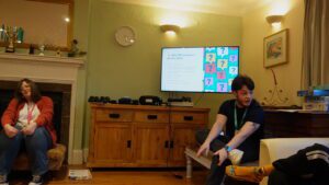 A man wearing a Meath staff lanyard talks to a man in a living room. Behind him is a presentation on a TV screen.