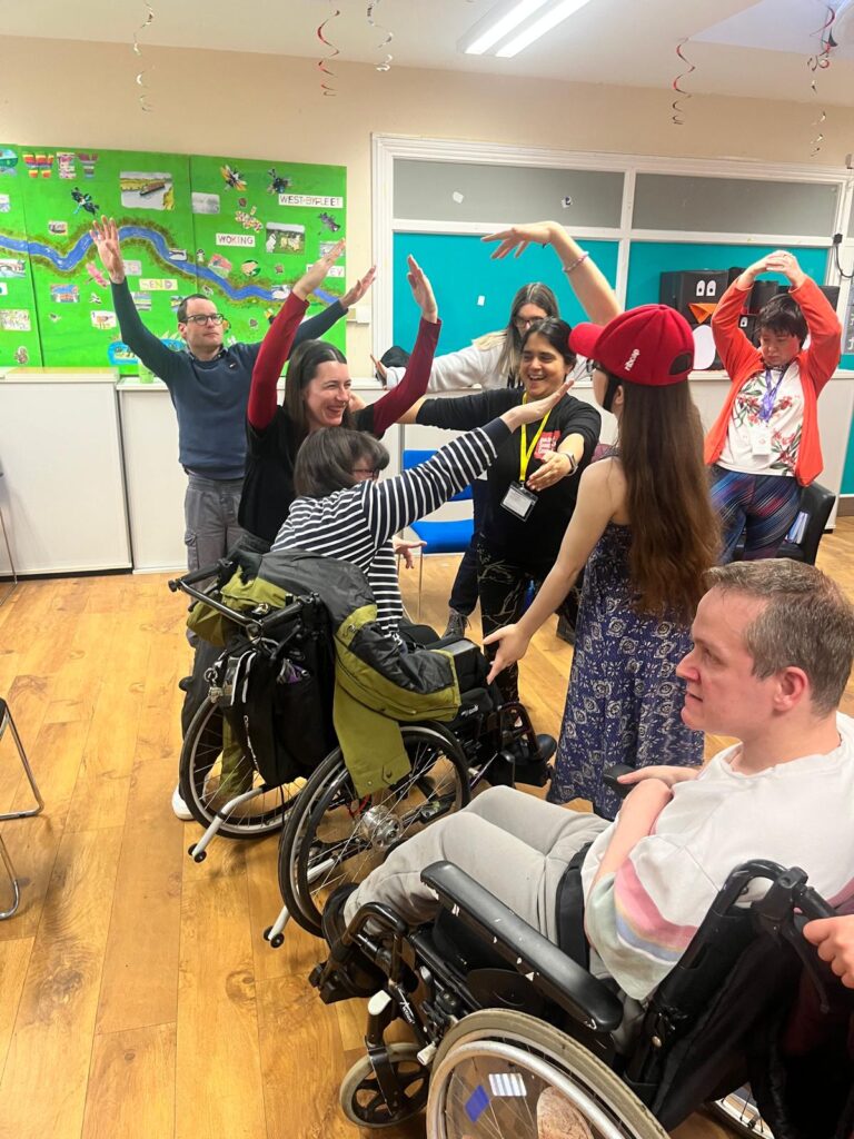 A group of men and women, some in wheelchairs are close together with their arms in the air in a bright room with a wooden floor.