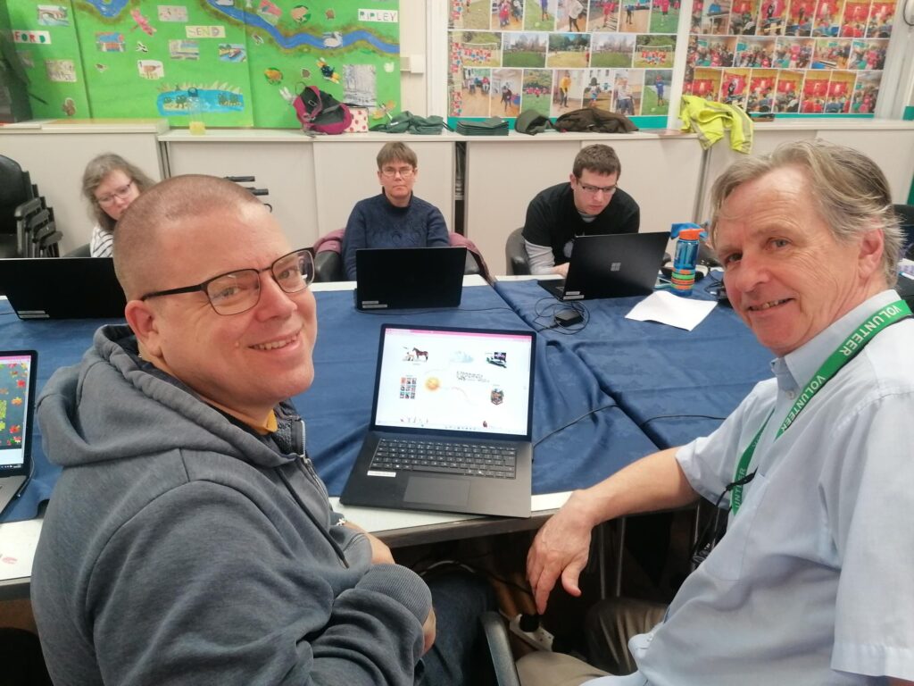 a man wearing a volunteers lanyard is helping a man to work at a lap top.
