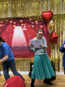 A young woman in teal tulle skirt and white top smiles as she holds a certificate. She has dark hair and glasses and is stood in front of a gold shimmer curtain.