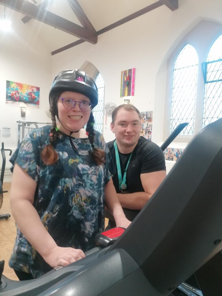 a woman wears an epilepsy helmet while using a treadmill. She smiles to the camera while a male staff member is stood next to her smiling.