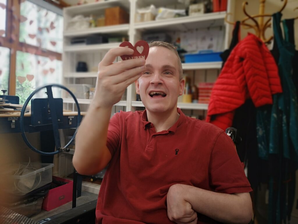 A man in a red top is smiling and holding up a small red wooden heart to the camera. He is in a wheelchair and is in a workshop.