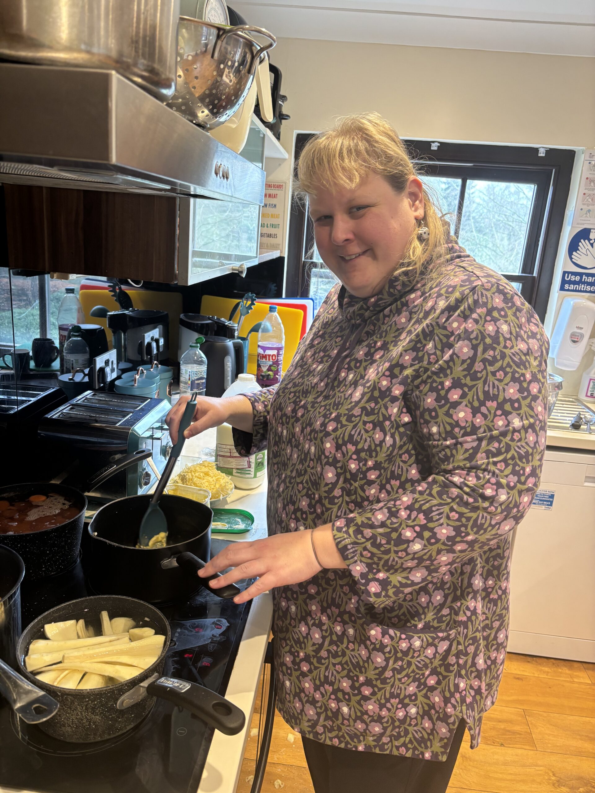 A woman in a floral dress is smiling as she cooks food in a pan on the hob in a kitchen.