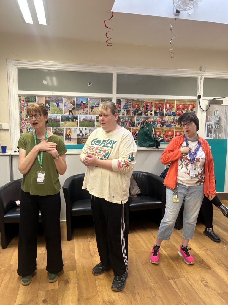 Three women stand in a large room with mouths open and hands signing. One woman wears a volunteer lanyard.