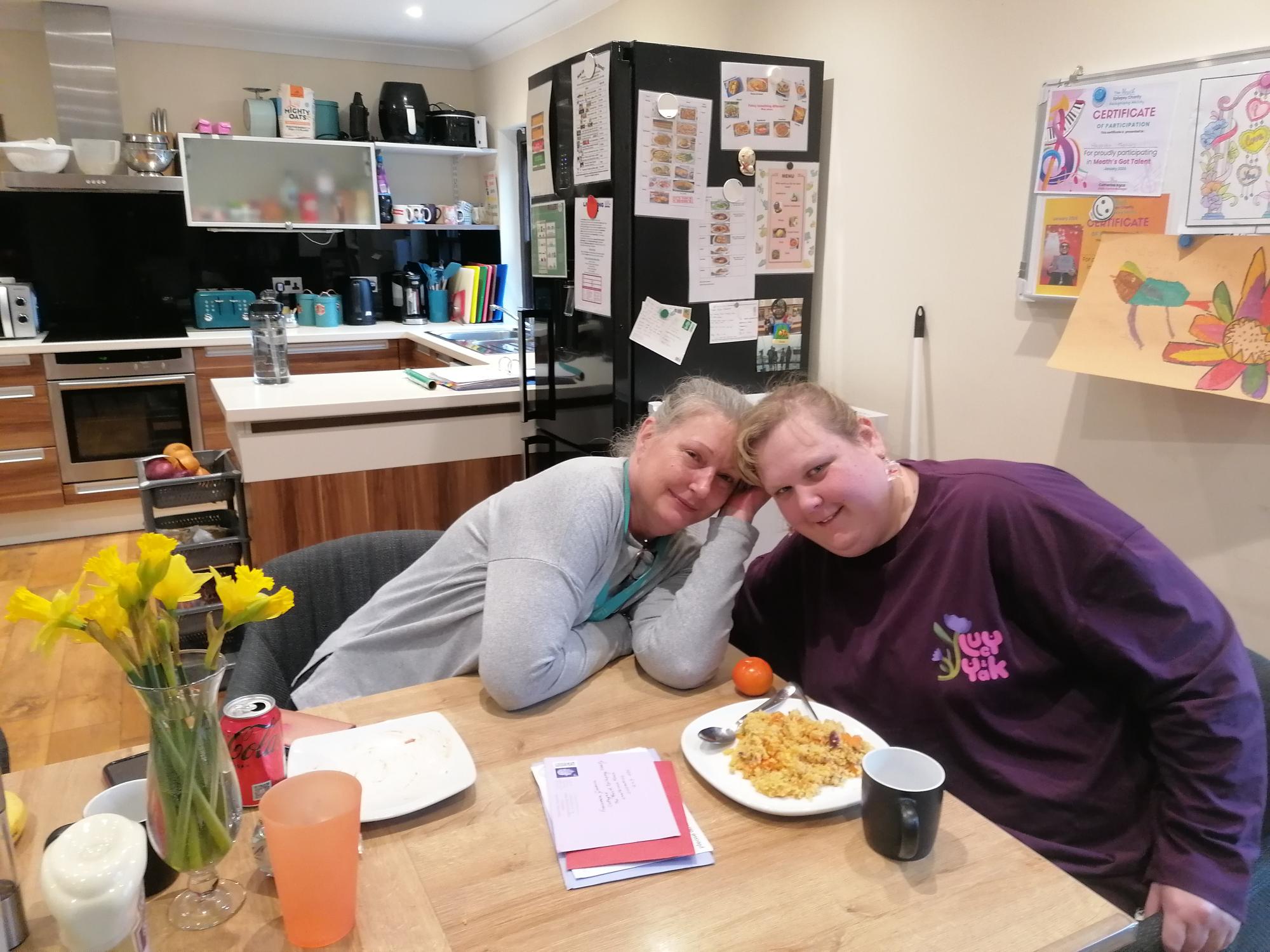 A young woman in a purple top is sat at her kitchen table with a colourful lunch in front of her. She is smiling and next to her is her support worker.