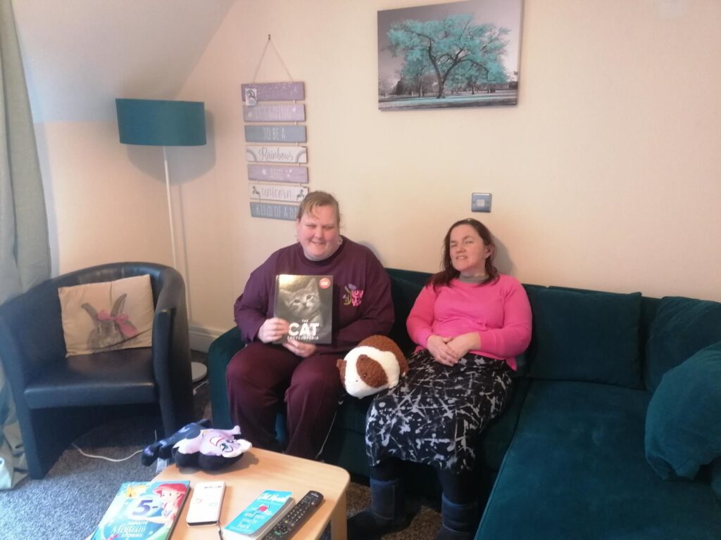 two women sit together in a living room on a teal sofa. Between them is a large cuddly Guinea Pig.
