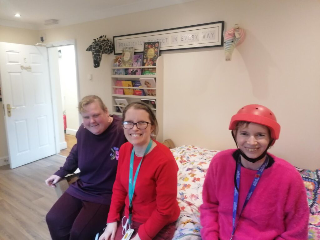 3 women smile at the cemera as they sit on a cosy bedroom together. One woman wears a red epilepsy helmet.
