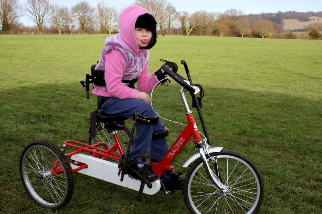 A girl in a pink top rides an adapted red bike. The bike has a harness.
