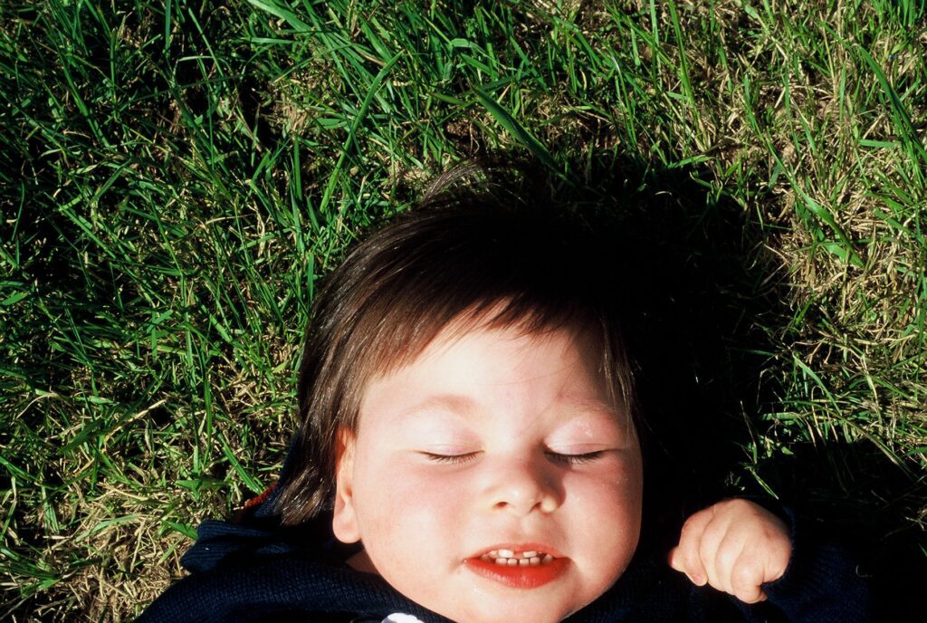 a young todlet with dark hair is pictured laying on green grass. She is smiling with her eyes closed.