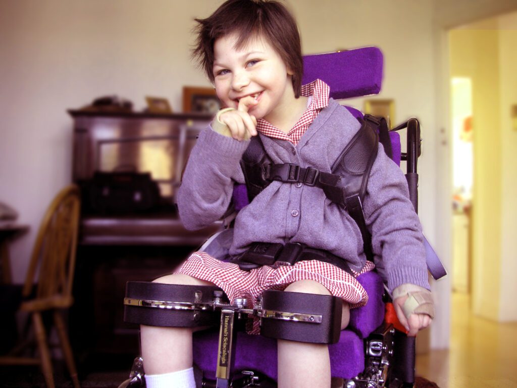 A girl wearing purple smiles to the camera. She is in a purple wheelchair and wears a harness.