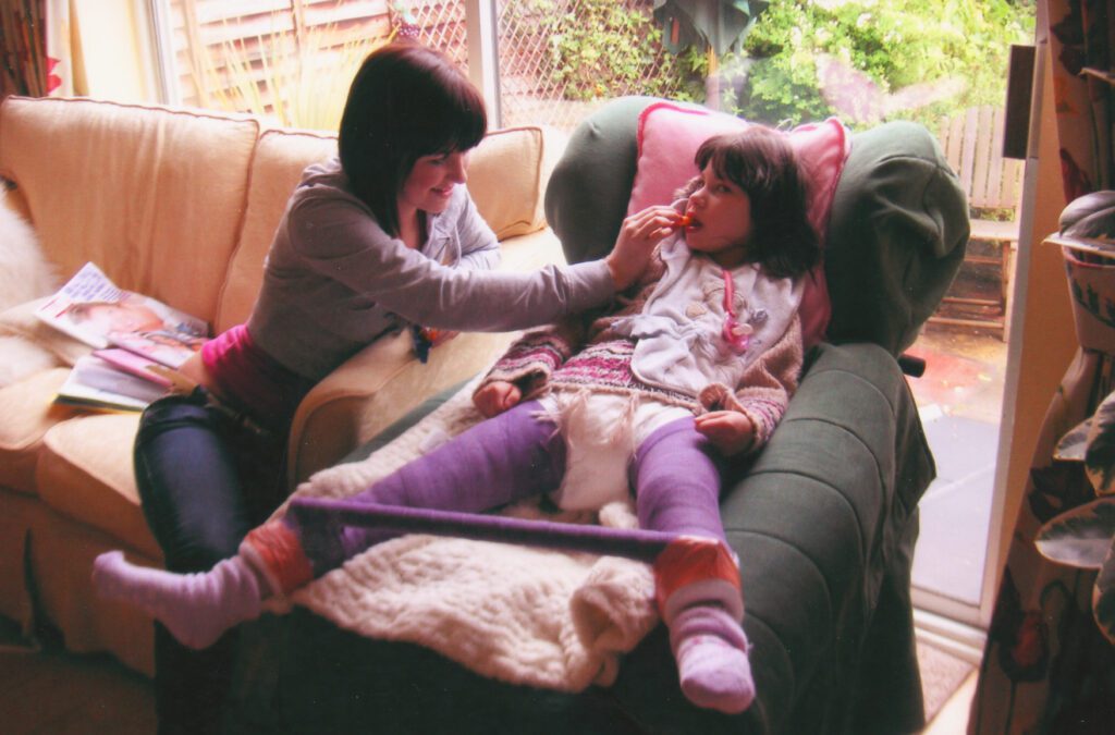 A young girl is pictured in an armchair next to her mother. The girl has her legs positioned in a post surgery a frame cast.