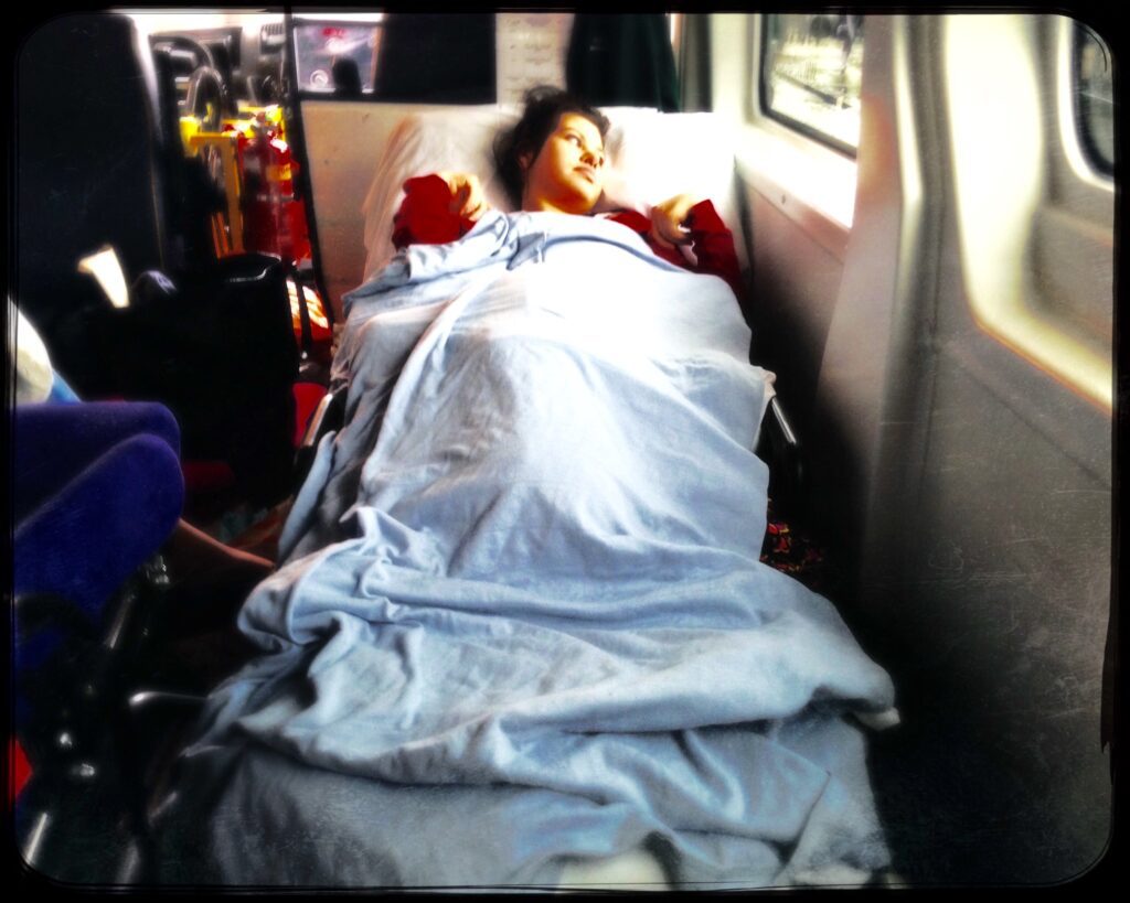 A girl with dark hair lies in an ambulance bed under a white sheet.