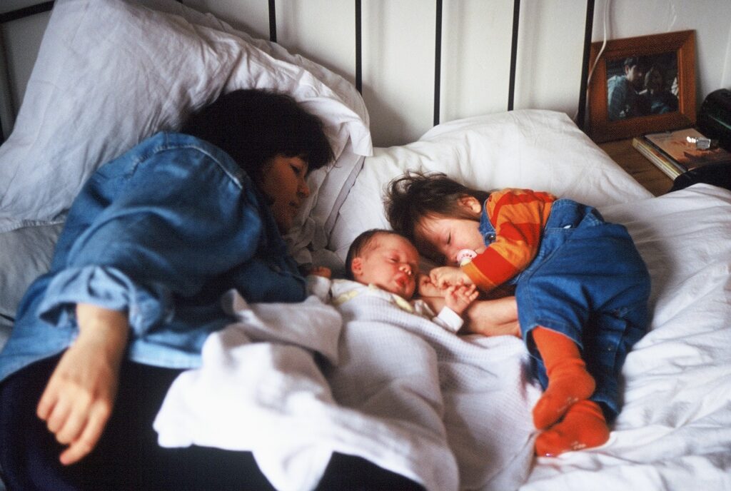 A woman lies on a bed, napping with a young baby and a toddler.