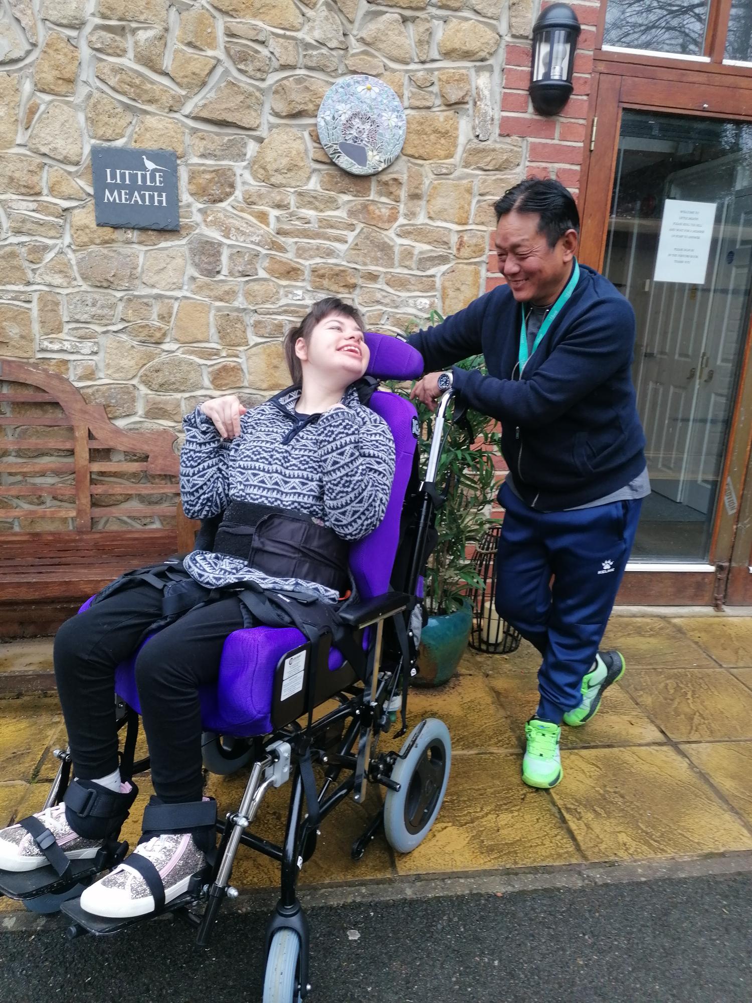 A young woman in a purple wheelchair wears a blue top and is looking up and smiling at a man. The man is wearing a Meath Epilepsy Charity lanyard.
