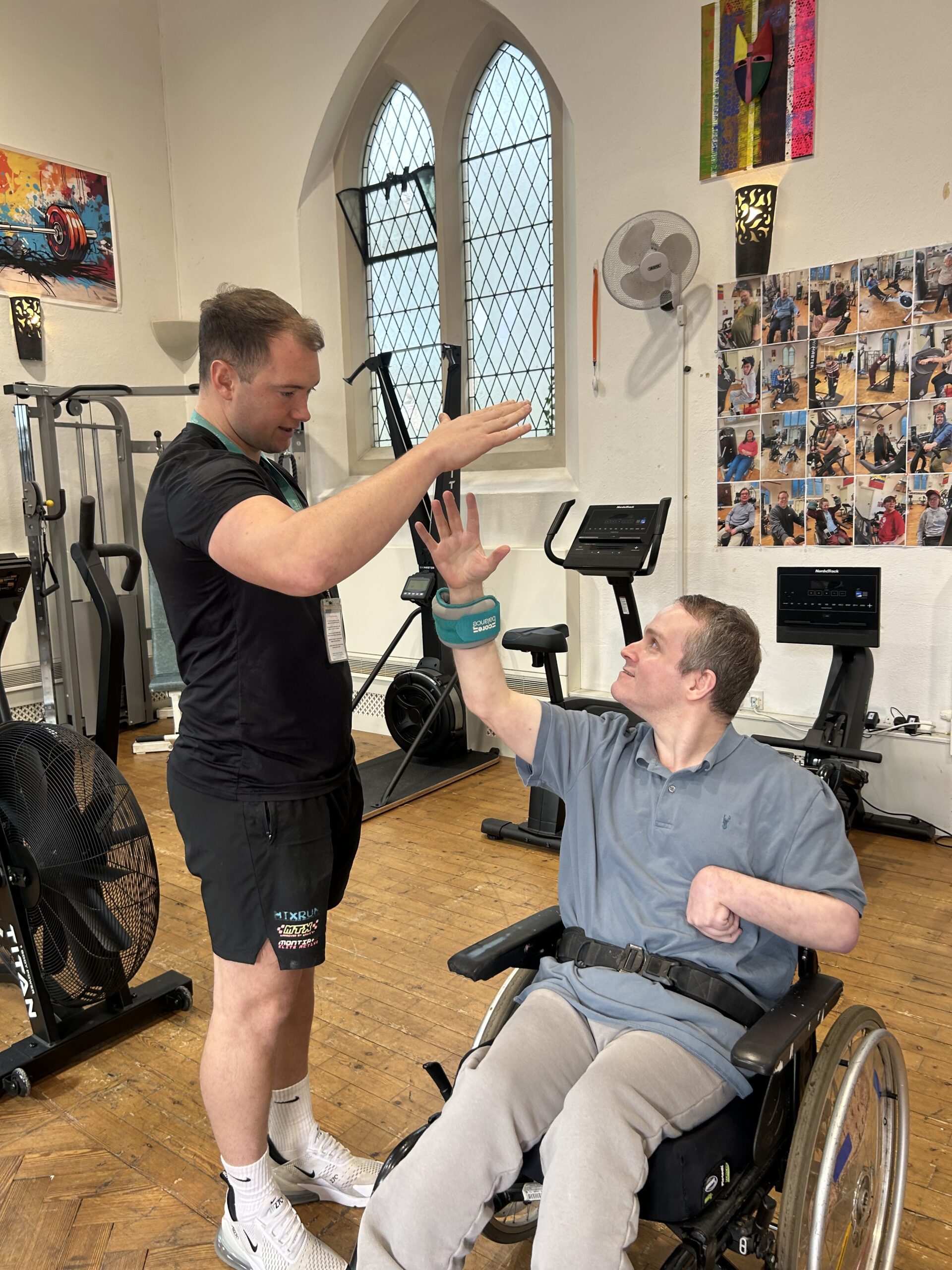 A man in a wheelchair is in a gym. He reaches out to high five a man who is stood next to him.