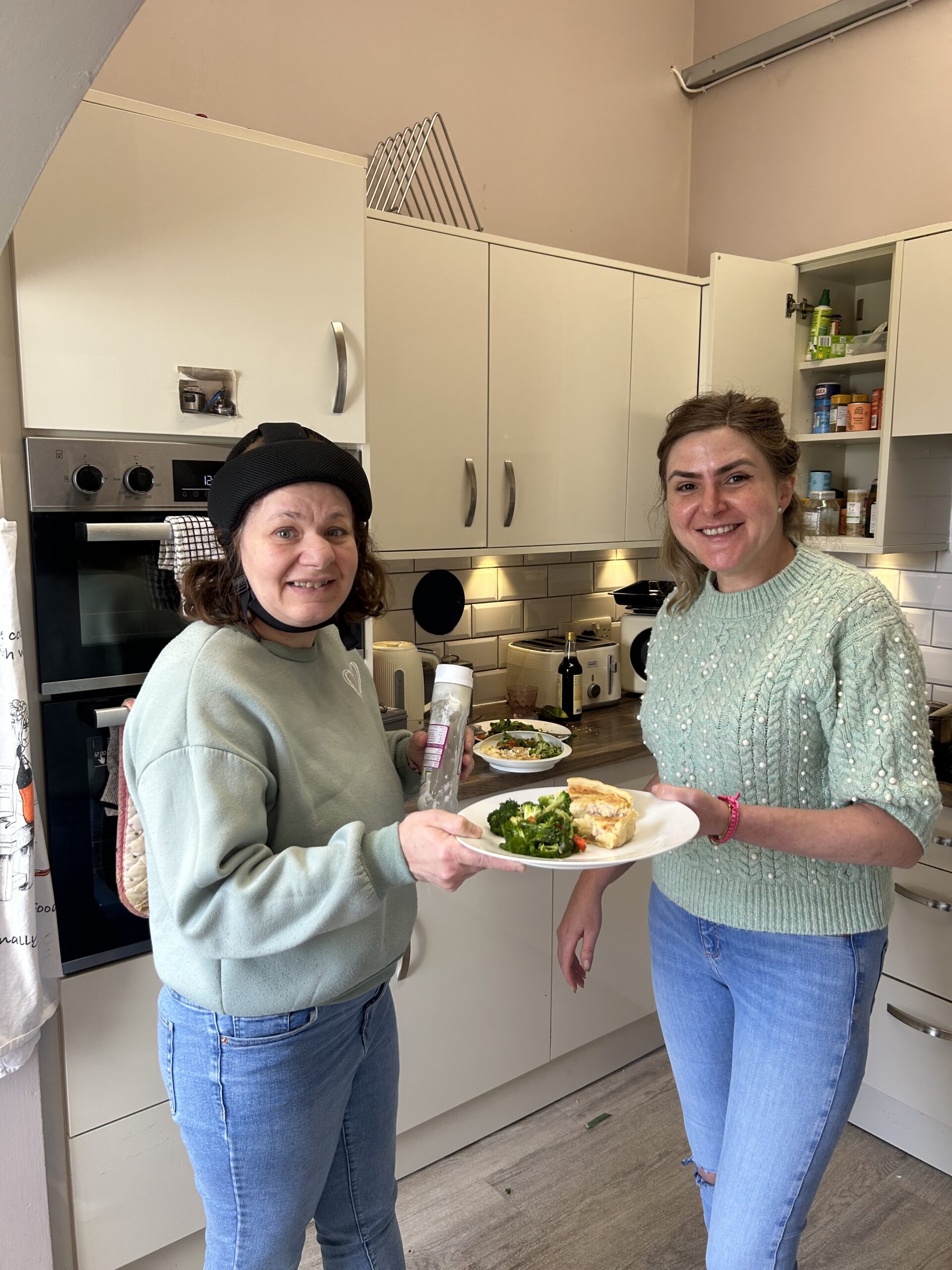 Two women smile in a kitchen as together they hold a plate. The plate has quiche and broccoli on it. One woman wears an epilepsy helmet. Both women wear blue jeans a pale green long sleeved top, they stand either side of the plate they are holding. The visual symetry and colour matching hints at the fact that they work as a team, despite one being a staff member and one being a person supported at The Meath Epilepsy Charity.