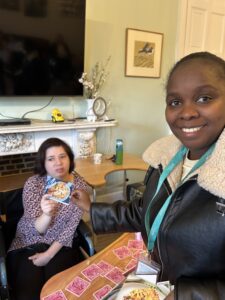 A young woman holds up a picture of a quiche with salad. Next to her is a smiling woman who wears a teal Meath lanyard. They are in a cosy living room interior.