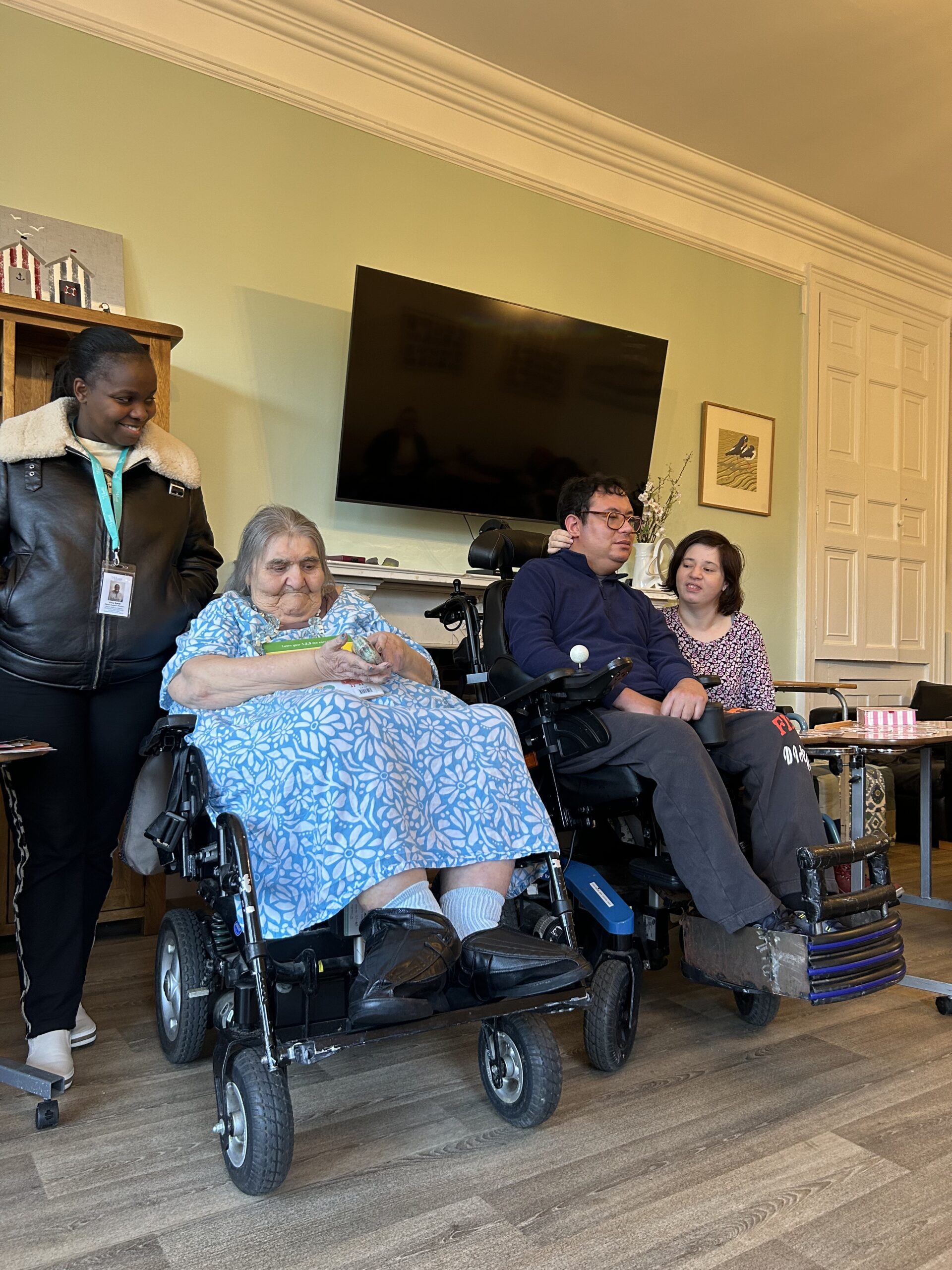 A woman in a blue dress sits in her wheelchair smiling in her home. She is next to her Support Worker.