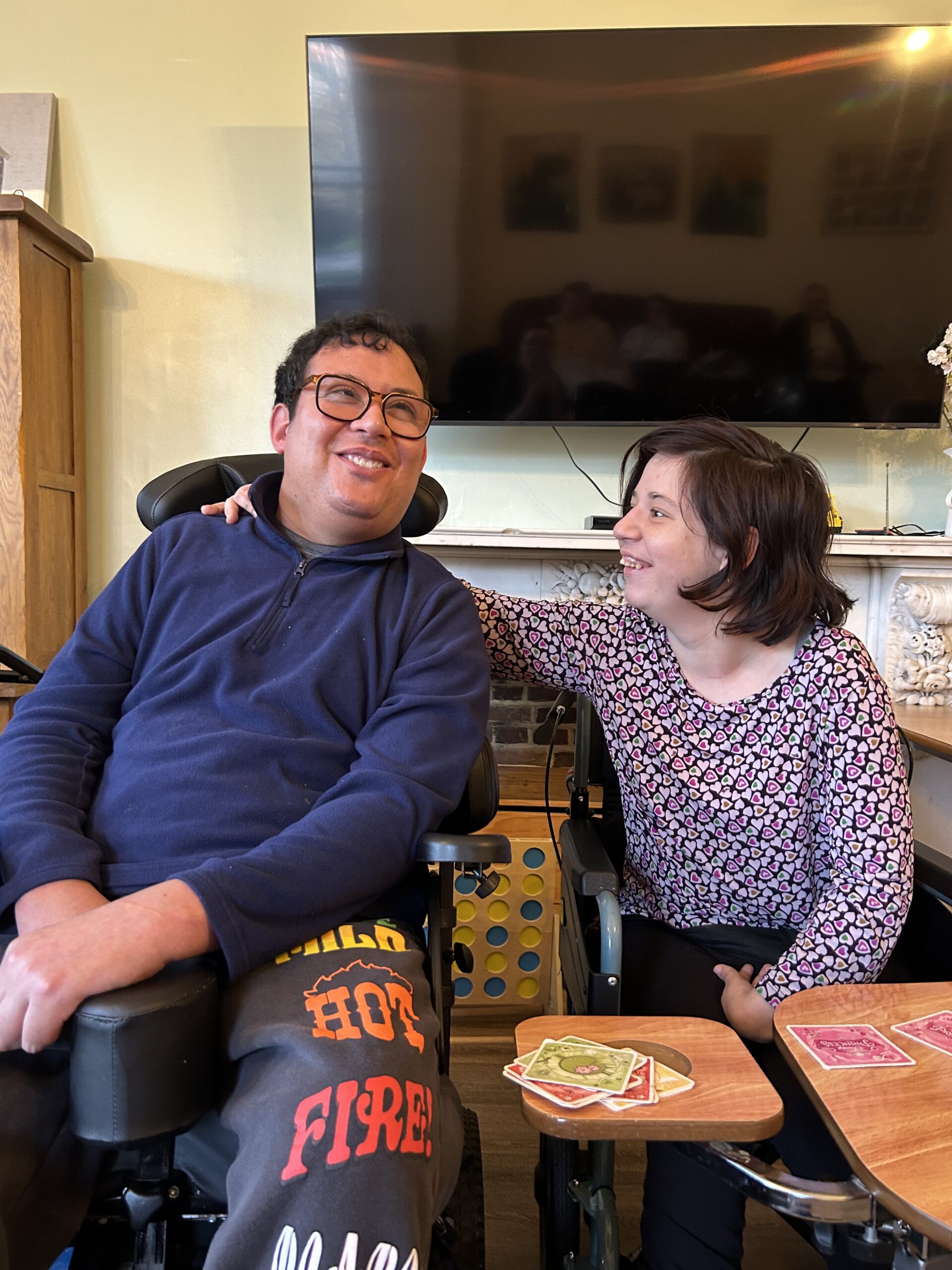 A young man with dark hair and glasses smiles and looks towards a young woman with dark hair who is smiling at him. They are sat next to each other in their wheelchairs. In front of them are cards laid out as they play a game.