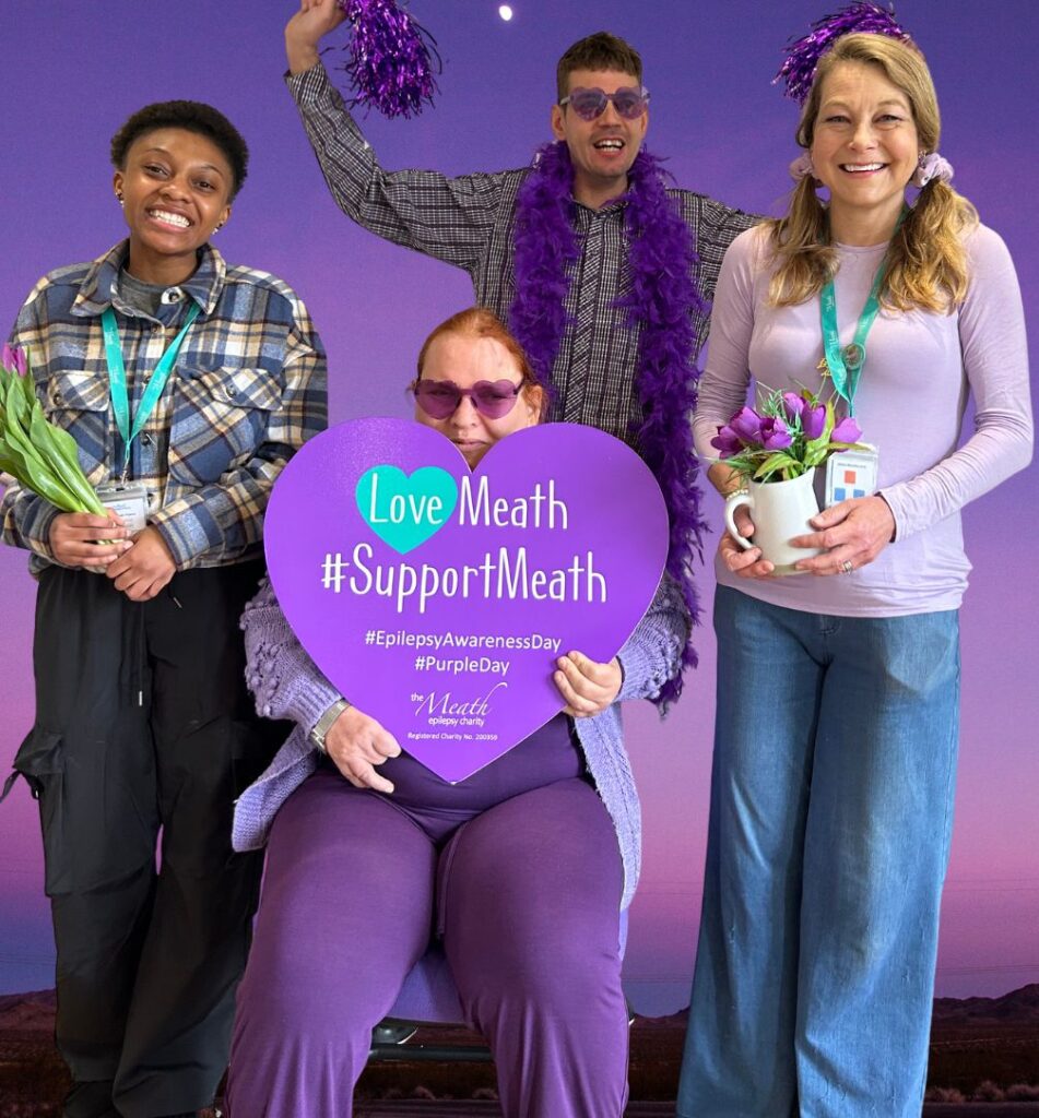 Four people are wearing purple clothes and accessories. They stand against a purple background smiling. One woman is seated and holds a large purple heart.