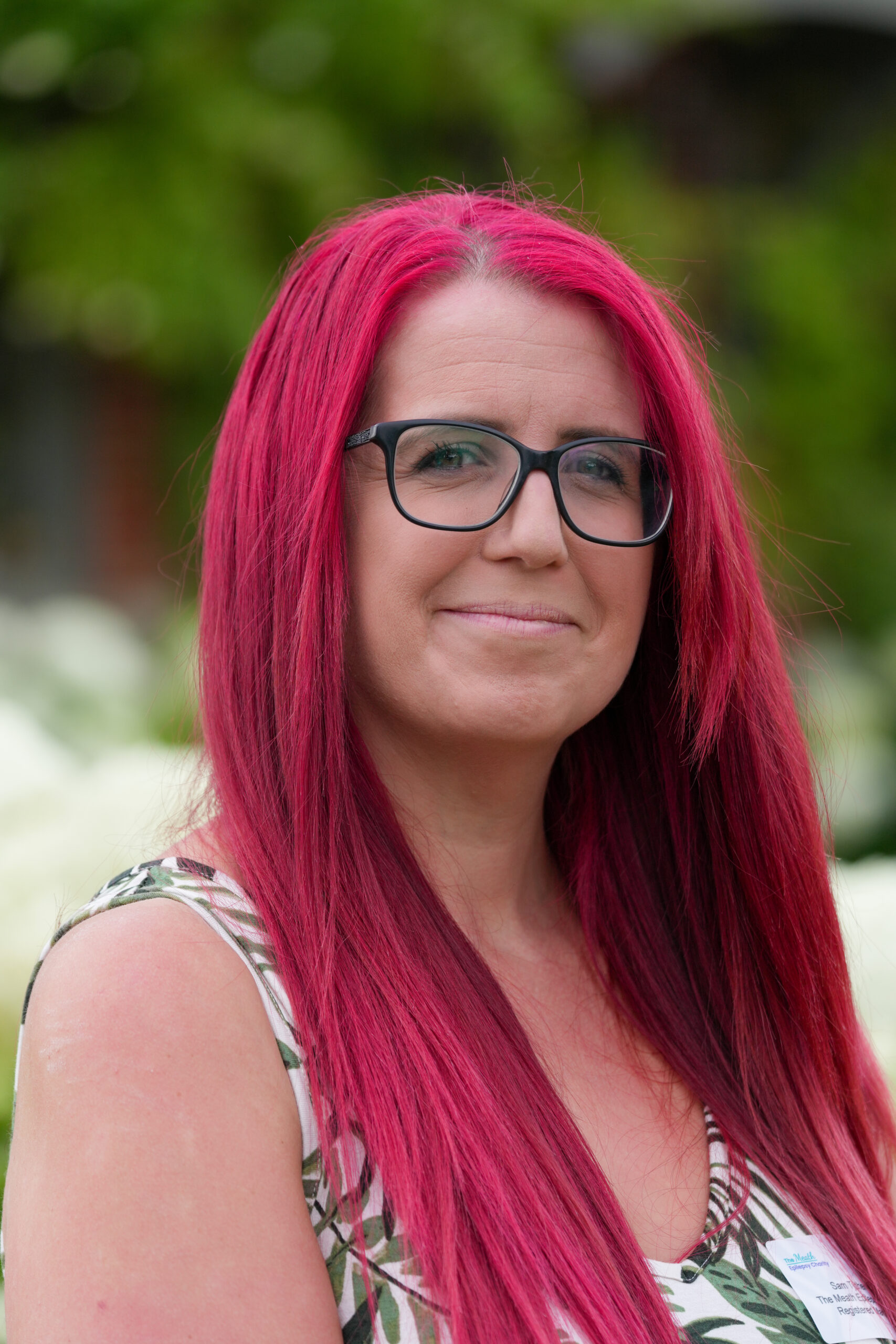 A woman with cherry red hair and dark framed glasses smiles at the camera. She stands against a backdrop of a garden in summer time.