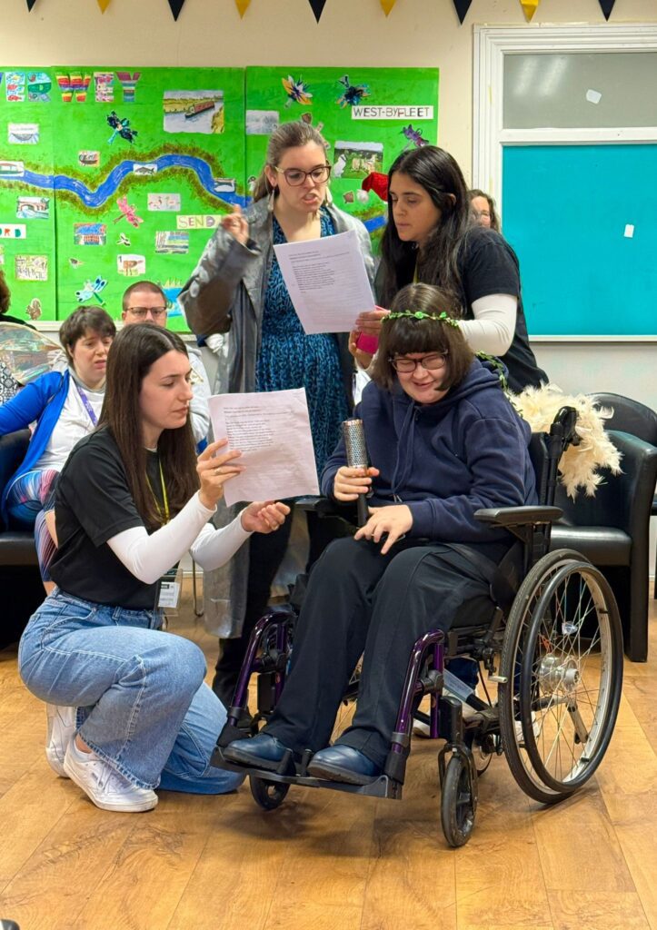 Four women with dark hair are together looking at scripts. One woman is in a wheelchair, another stands wearing a silver jacket and holding a wand.