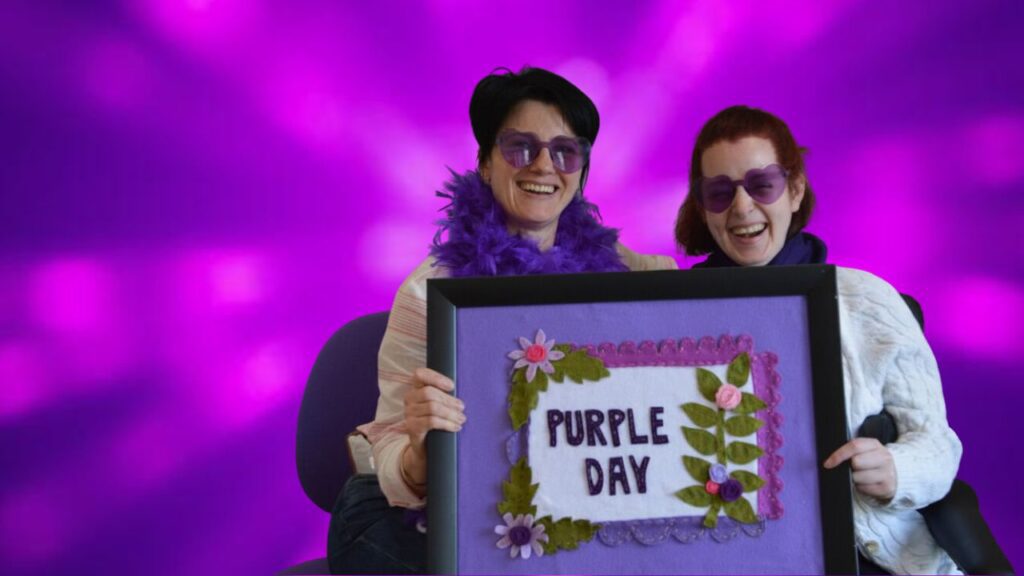 Two women smile as they wear purple heart glasses and hold a sign saying 'Purple Day'. They are seated in front of a bright purple background.