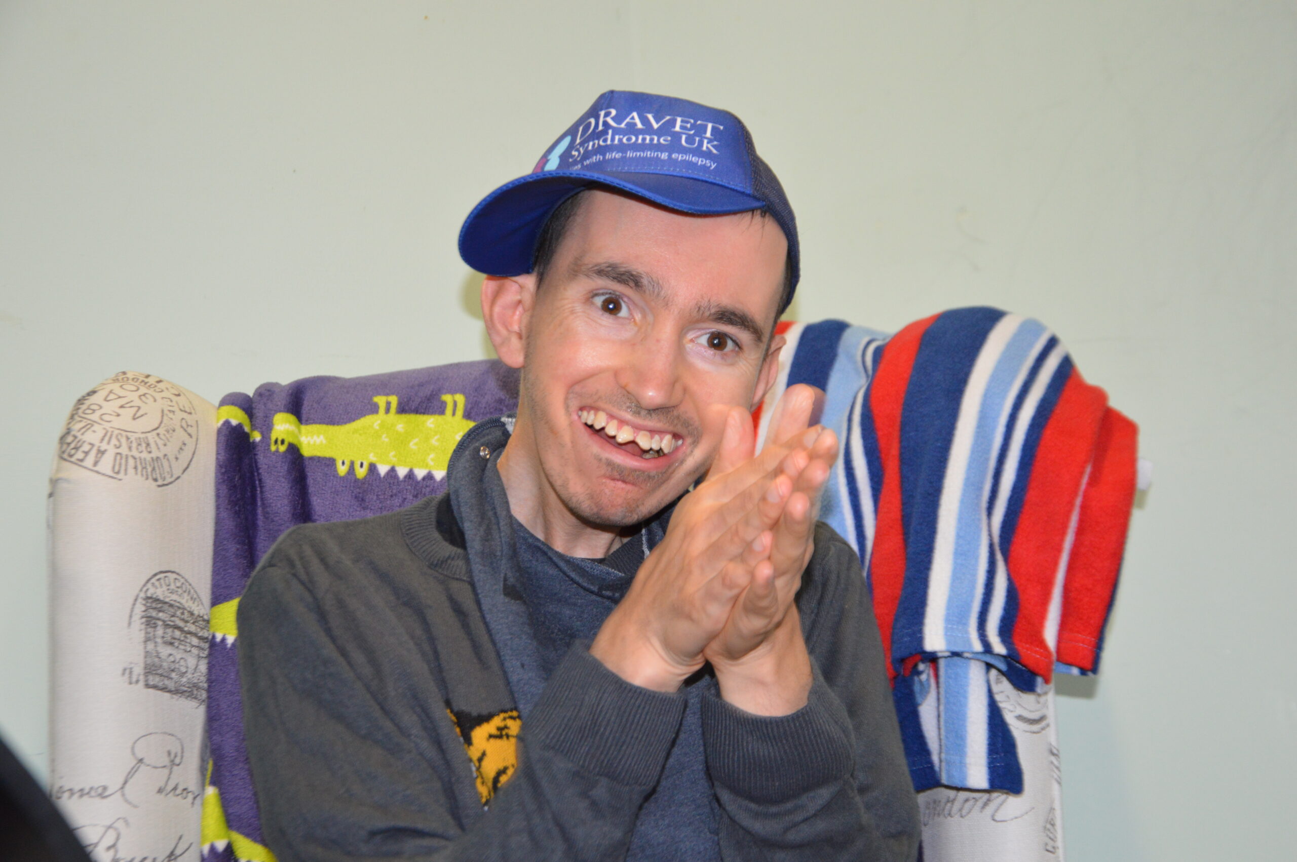 A man wearing a purple cap with 'Dravet Syndrome' on it sits in an arm chair and claps as he smiles to the camera. Behind him is a colourful blanket.