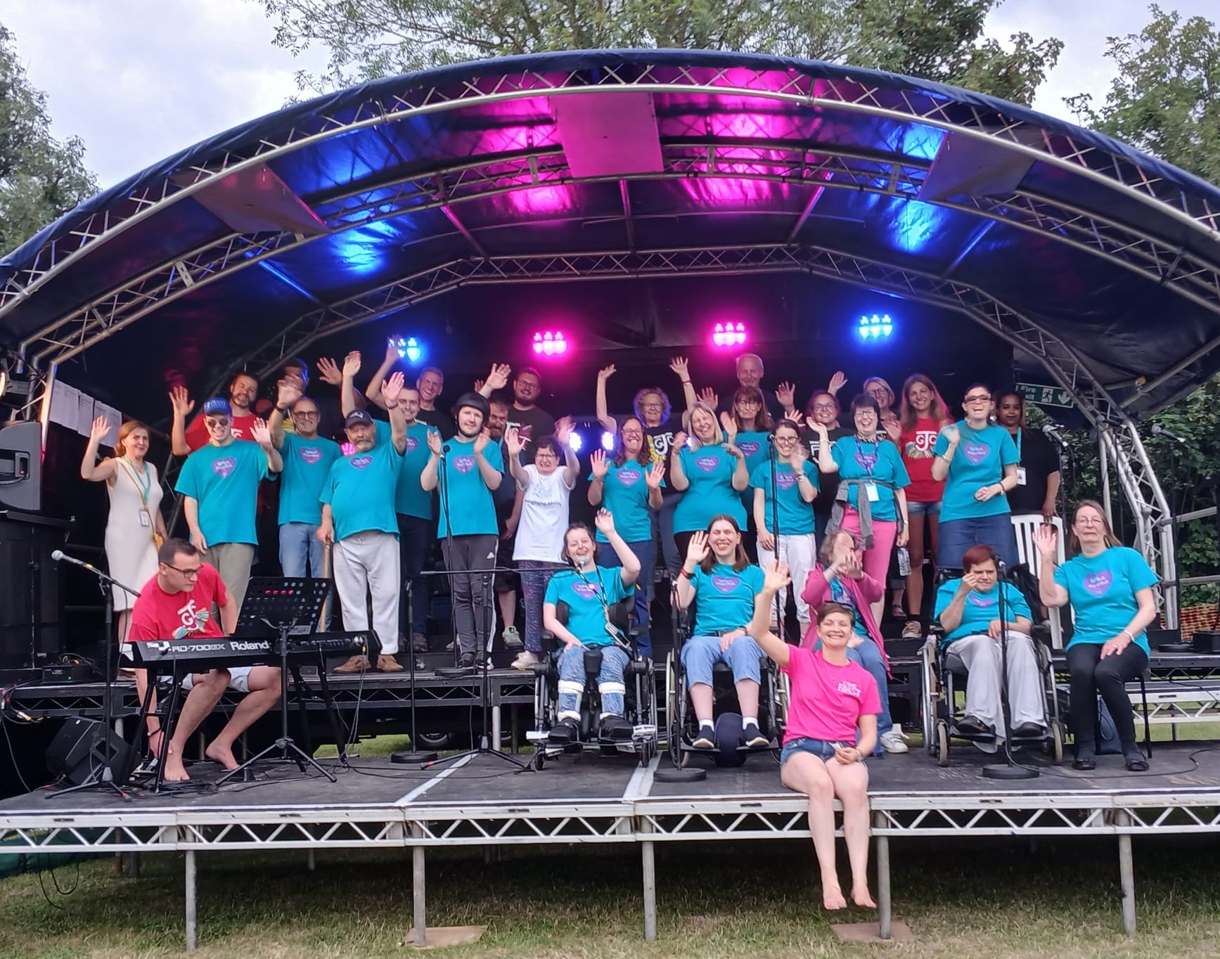 The Meath choir waving on an outside stage. They are wearing teal Meath T-shirts.