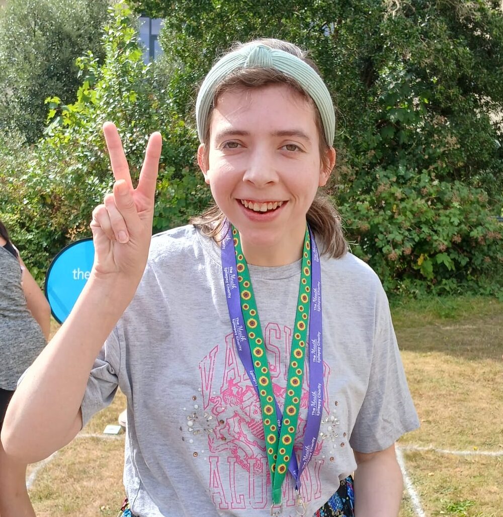 A woman stands on a large lawn and smiles while giving a peace sign and holding an egg and spoon. In the background are other race participants and a staff member supporting someone to take part.