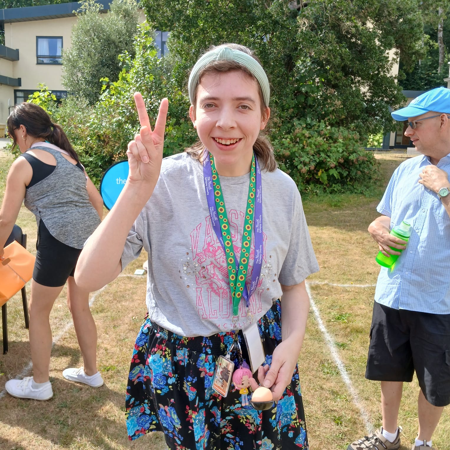 A woman stands on a large lawn and smiles while giving a peace sign and holding an egg and spoon. In the background are other race participants and a staff member supporting someone to take part.