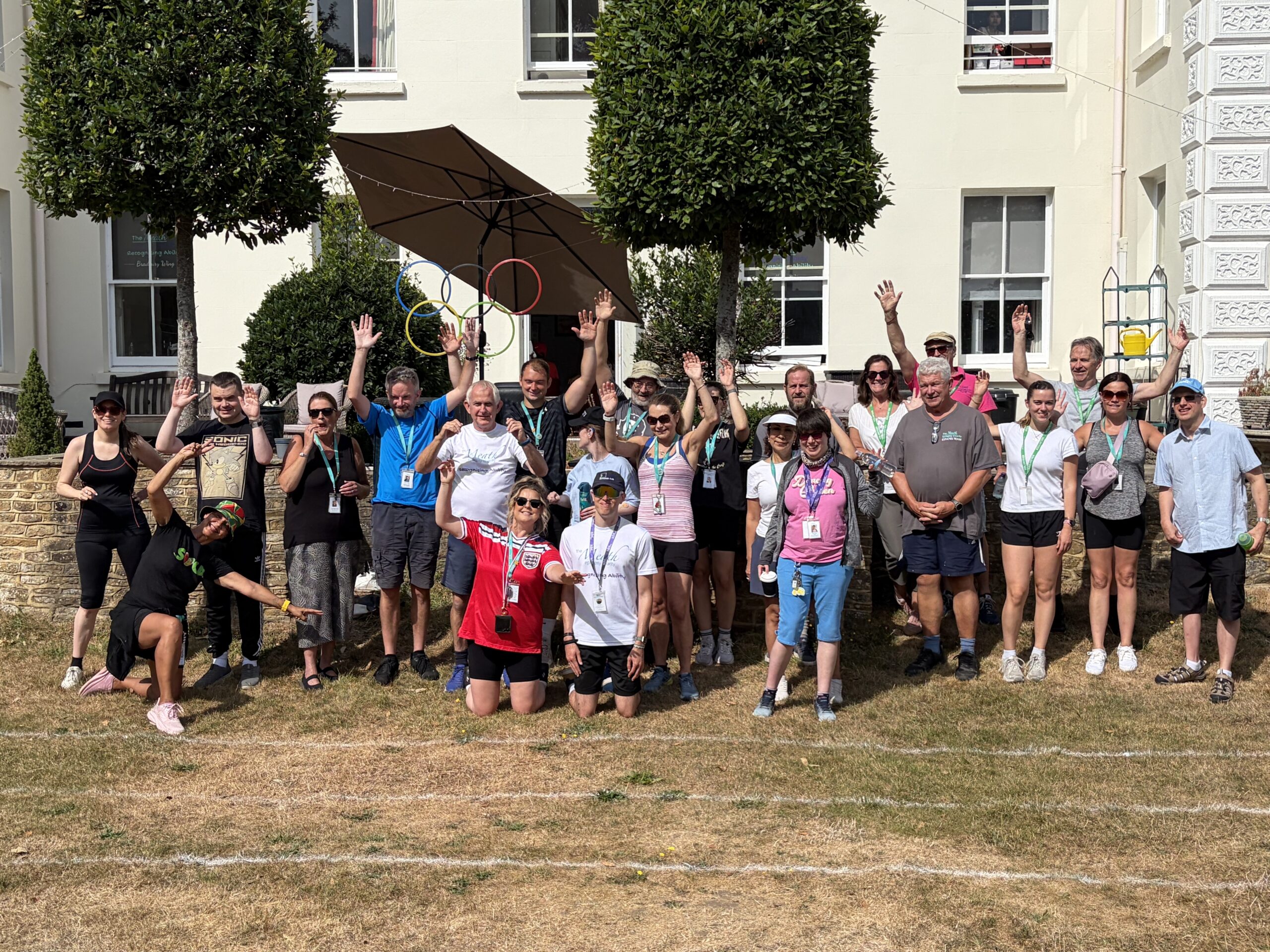 A big group of people stand in a crowd and cheer. They are wearing colourful sports wear and are stood/kneeling in fron of a large white Manor House on a lawn in summer.